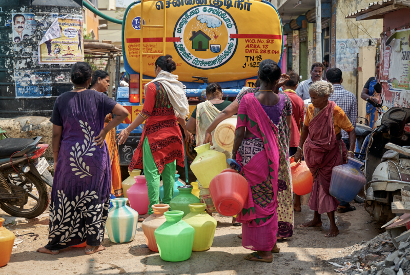 About six women wearing colorful saris stand behind a water tanker truck on a busy city street, waiting to fill buckets they are holding