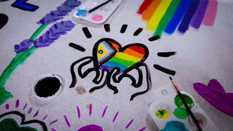 A close-up of a painted mural. On a white background, students have hand-painted lavender stems, a rainbow, and hearts in various colors. The design in the center of the photo shows the outlines of two people holding up a heart painted with the pride flag design. A paintbrush and a container with several colors of paint in it sits in the bottom right corner of the photo.